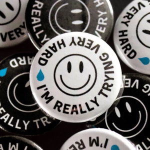 May include: A collection of round buttons with a smiley face design. The buttons are white and black, with text that reads "I'm Really Trying Very Hard." Some buttons have a blue teardrop graphic. The buttons are a fun, quirky accessory.