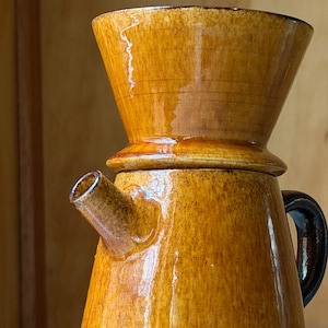 Op de afbeelding: Een bruine en tan keramische pour-over koffiemaker. De koffiemaker heeft een conische bovenkant, een tuit en een handvat. Hij staat op een bijpassende schotel. Het ontwerp is geglazuurd met een glanzende afwerking.