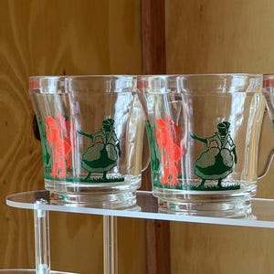 May include: Clear glass mugs with a handle, featuring a green and black illustration of a person in a dress. The design also includes a red figure. The mugs are displayed on a clear acrylic shelf.