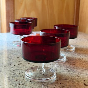 Può includere: Un set di cinque coppette da dessert in vetro rosso rubino, ciascuna caratterizzata da un'ampia coppa cilindrica sostenuta da un gambo corto in vetro trasparente e una base circolare. Le coppette hanno una finitura liscia e uniforme.