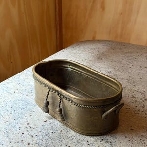 Puede incluir: Una jardinera de latón antiguo con forma ovalada. La jardinera presenta un diseño de cuerda alrededor del borde y dos pequeñas borlas. Tiene dos asas pequeñas en los laterales.