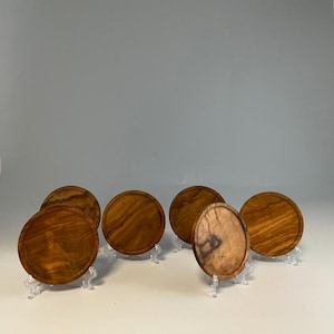 May include: A set of six circular wooden coasters featuring a distinct natural grain pattern in shades of light and dark brown. Each coaster is displayed upright on a small clear plastic stand, showcasing the smooth finish and circular shape.