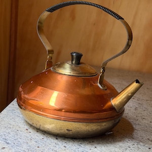 May include: A tea kettle featuring a polished copper upper body and a brass lower base. It includes a brass lid with a black knob, a brass spout, and a curved metal handle with a black textured grip. The design is rounded with a wide base.