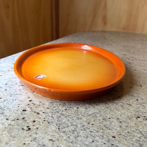 May include: An orange and yellow ceramic plate with a rounded edge. The plate has a glossy finish and is sitting on a speckled countertop. The plate is round and has a small sticker on the inside.
