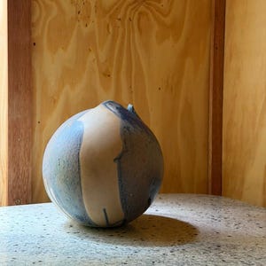 Könnte beinhalten: Eine kugelförmige Keramikvase mit einer glatten, matten Oberfläche. Das Design zeigt eine Mischung aus natürlichen Beigetönen und gesprenkelten blau-grauen Flecken, die einen deutlichen Farbkontrast auf der runden Form des Gefäßes erzeugen.