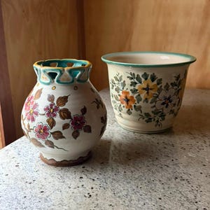Op de afbeelding: Een keramieken vaas met een geschulpte blauwgroene rand en handgeschilderde roze bloemmotieven naast een bijpassende keramieken plantenbak met een brede blauwgroen omrande opening en een geschilderd patroon van gele, oranje en witte bloemen met groene bladeren.