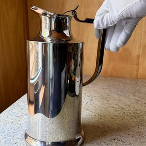 Könnte beinhalten: Eine weiße Keramik-Tasse mit schwarzem Henkel und dem Text "But First, Coffee" in schwarzer Schrift. Die Tasse ist mit einer dunklen Flüssigkeit, wahrscheinlich Kaffee, gefüllt und steht auf einer hellen Oberfläche.