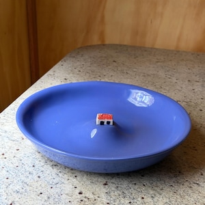 Puede incluir: Un cuenco de cerámica azul bígaro con una pequeña y detallada casa en miniatura en el centro. La casa tiene un tejado rojo y paredes blancas. El cuenco está sobre una encimera moteada.