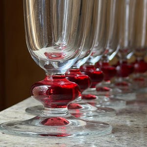 Könnte beinhalten: Eine Reihe klarer Champagnerflöten mit einem leuchtend roten Fuß und Stiel. Die Flöten sind in einer Reihe angeordnet und zeigen ihr elegantes Design und die satte Farbe des Fußes. Die Gläser stehen auf einer gesprenkelten Oberfläche.