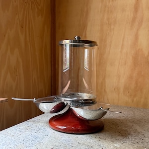 May include: A condiment server featuring a central clear glass cylindrical container with a silver-toned metal lid, surrounded by three attached silver-toned metal bowls with matching spoons, all mounted on a circular dark reddish-brown wooden base.