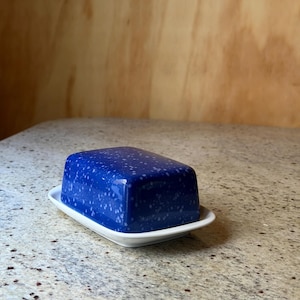 Op de afbeelding: Een blauwe keramische botervloot met een gespikkeld patroon, rustend op een witte rechthoekige basis. De schaal is rechthoekig met afgeronde randen. De achtergrond is een lichtbruin houten oppervlak.