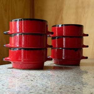 May include: Six red ceramic bowls with black trim are stacked in two groups of three. Each bowl has two small handles. The bowls are on a speckled countertop with a wooden background.