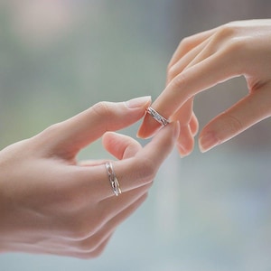 Può includere: Vengono mostrate due fedi d'argento, una delle quali viene posta su un dito. Gli anelli hanno disegni intricati. Lo sfondo è sfocato, con un focus sulle mani e sugli anelli. Gli anelli sembrano essere per una coppia.