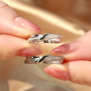 May include: Two silver-toned rings with a crossover design. The top ring features a band encrusted with small clear stones, while the bottom ring has a smooth, polished finish. Both rings exhibit a curved, overlapping structure in a metallic material.