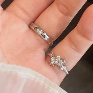 Peut inclure: Deux bagues en argent aux motifs complexes. L'une présente un motif en étoile et un petit diamant, l'autre un motif floral et étoilé avec plusieurs petits diamants. Les bagues sont présentées sur une main.