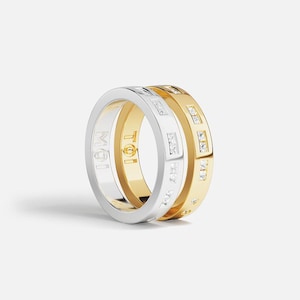 Puede incluir: Dos anillos apilados, uno plateado y otro dorado. El anillo plateado tiene la inscripción "Moi", y el dorado "Toi". Ambos anillos tienen pequeñas gemas transparentes. Una joya elegante.
