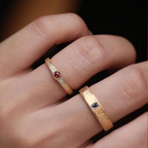 May include: Two gold rings with textured bands. One ring features a small, round red gemstone, while the other has a rectangular blue gemstone. The rings are displayed on a hand.
