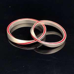 May include: Two silver rings with a red enamel stripe. The rings are intertwined, creating a unique design. The rings are displayed on a reflective black surface.