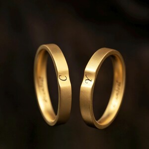 May include: Two gold-toned rings with a matte finish, each featuring a subtle V-shaped indentation on the outer band. One ring is engraved with the letter C, and the other ring is engraved with the letter Y. Both bands show additional text inside.
