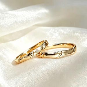 Op de afbeelding: Twee gouden ringen met een gepolijste afwerking. De ene ring is voorzien van een reeks kleine, heldere, rondgeslepen stenen in de band, terwijl de andere ring een glad, effen gouden oppervlak heeft met subtiele, gelijkmatige inkepingen.
