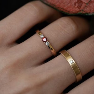 May include: Two gold rings. One ring features a central red gemstone flanked by two smaller clear gemstones. The other ring is a simple gold band with a single, small, clear gemstone. The rings are on a finger.