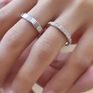 Op de afbeelding: Twee zilveren ringen. Eén ring heeft een reeks vierkante segmenten. De andere ring is versierd met kleine, vierkante edelstenen. Beide ringen zitten aan de vingers van een persoon.