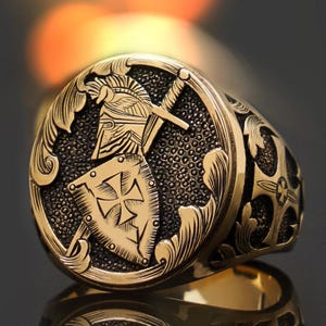 Puede incluir: Un anillo dorado con un diseño detallado de un yelmo de caballero, una espada y un escudo con una cruz. El anillo tiene una superficie ornamentada y texturizada, con un acabado pulido, mostrando un tema histórico clásico.