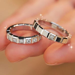 Op de afbeelding: Twee zilveren ringen met een geometrisch ontwerp. Elke ring heeft vierkant geslepen edelstenen, gezet in een reeks rechthoekige frames. De ringen worden op een hand getoond, met een zachte, vage achtergrond.