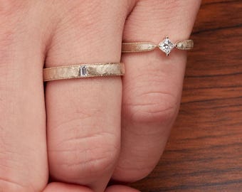 May include: Two gold rings with textured bands. One ring features a rectangular gemstone, while the other showcases a square-cut diamond. The rings are displayed on a wooden surface, highlighting their intricate design and craftsmanship.