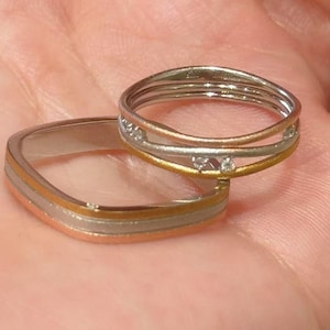 May include: Two rings with a mix of silver, gold, and rose gold tones. One ring is square with three bands, and the other is a stack of three rings with small gemstones.