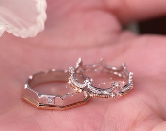 Puede incluir: Se muestran dos anillos, uno con diseño de corona y otro con forma geométrica. El anillo de corona es plateado con pequeñas piedras transparentes. El otro anillo es plateado y dorado con una pequeña piedra cuadrada.