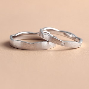 May include: Two silver rings with a wavy design. Each ring has a brushed metal finish and a small, clear gemstone. The rings are set against a neutral background, highlighting their elegant simplicity. Ideal for couples.