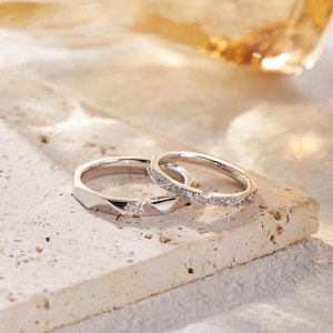 May include: Two silver rings with geometric designs. One ring features a series of small, sparkling stones along the top edge. The other ring has a faceted design with a single stone. The rings are displayed on a textured, light-colored surface.