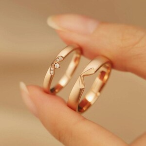 Op de afbeelding: Twee gouden trouwringen. Eén ring heeft een gebogen ontwerp met kleine, sprankelende stenen. De andere ring heeft een geometrisch, hoekig ontwerp. De ringen worden tussen de vingers gehouden, waarbij hun gepolijste afwerking en details worden benadrukt.