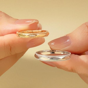 Könnte beinhalten: Zwei Ringe, einer goldfarben und einer silberfarben, werden gezeigt. Der goldfarbene Ring hat ein rautenförmiges Muster mit kleinen Diamanten in der Mitte. Der silberfarbene Ring hat ein ähnliches Design. Beide Ringe werden vor einem weichen, neutralen Hintergrund gehalten.