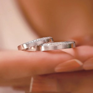 May include: Two silver rings with a brushed metal finish, each encrusted with small, sparkling gemstones along the top edge. The rings are displayed on a blurred background, suggesting a focus on the jewelry.