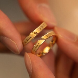 May include: Two gold rings, one broken, held in a person's hand. The rings have the words "IN THE CRACK" and "LIGHT" engraved on them. The rings are a warm gold color.
