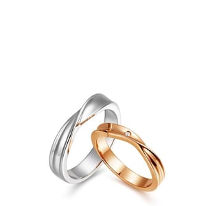 May include: Two intertwined rings, one silver and one rose gold, against a white background. The rose gold ring features a small, clear gemstone. The rings have a smooth, twisted design, symbolizing unity and connection. Ideal for weddings or anniversaries.