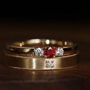 Può includere: Due anelli d'oro, uno con una gemma rossa centrale affiancata da due gemme bianche, e l'altro con una singola gemma bianca. Gli anelli sono appoggiati su una superficie di legno scuro.