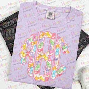 May include: A purple t-shirt with a pink outline of the letters "RY" filled with a pattern of pastel-coloured Peeps candy. The Peeps are arranged in a random pattern and include pink, yellow, blue, and green colours.