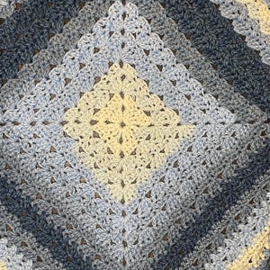 Puede incluir: Una manta de ganchillo hecha a mano con un patrón cuadrado. La manta presenta un degradado de colores, comenzando con un centro crema claro y pasando por tonos de azul y azul marino. Las puntadas de ganchillo crean una apariencia texturizada y acogedora.