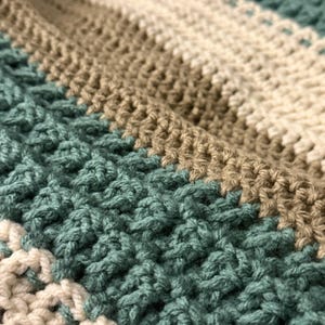 Könnte beinhalten: Nahaufnahme einer handgefertigten Häkeldecke. Die Decke hat abwechselnde Streifen aus Creme, Beige und Türkis. Die Textur des Häkelstichs ist sichtbar und erzeugt ein gemütliches und strukturiertes Aussehen. Die Decke ist wahrscheinlich aus weichem Garn gefertigt.
