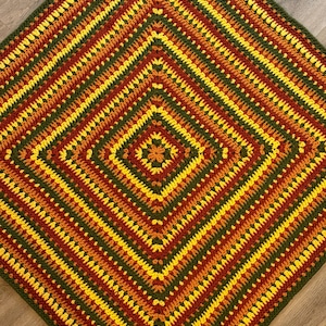 May include: A handmade crochet blanket with a geometric diamond pattern. The blanket features concentric squares in shades of green, yellow, orange, and brown. The crochet stitches are visible, creating a textured surface. The blanket is likely used for home decor.