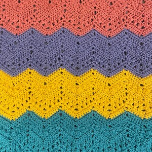 May include: A close-up of a crocheted blanket with a chevron pattern. The blanket features bands of color in coral, lavender, yellow, and teal. The crochet stitches create a textured surface with small openings throughout the design.