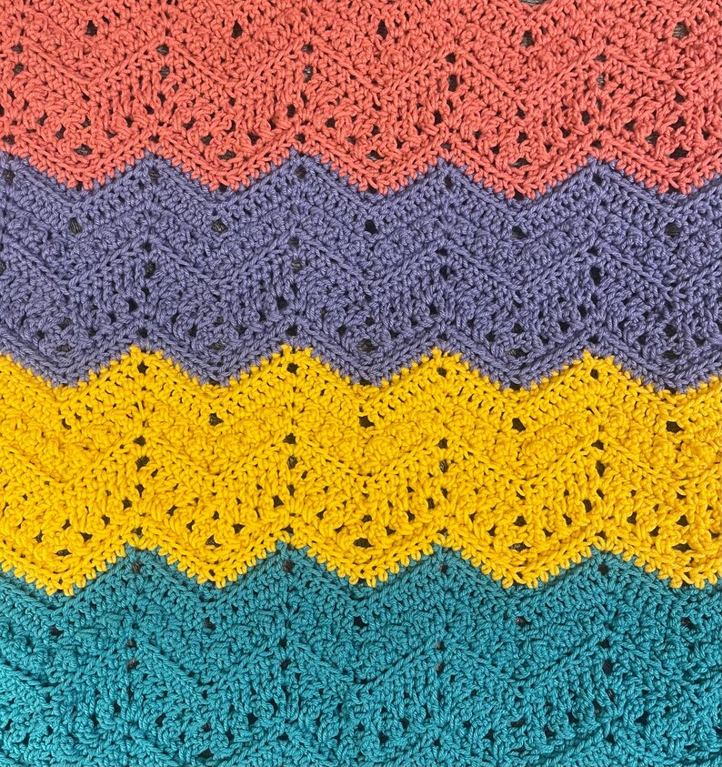 May include: A close-up of a crocheted blanket with a chevron pattern. The blanket features bands of color in coral, lavender, yellow, and teal. The crochet stitches create a textured surface with small openings throughout the design.