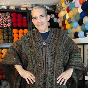 Puede incluir: Un poncho de ganchillo hecho a mano, marrón, verde y azul, con un diseño de cuello en V. El poncho tiene un patrón texturizado y lo lleva una persona. El fondo muestra estantes llenos de hilo de varios colores.