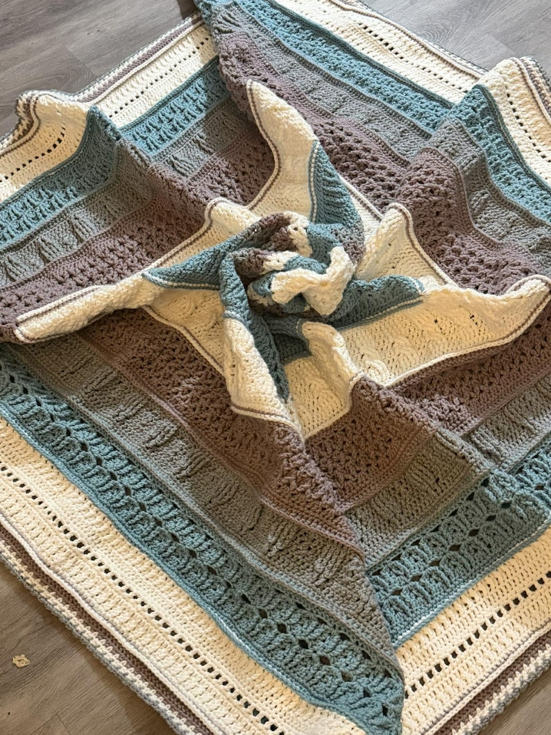 May include: A square crochet blanket featuring a repeating pattern of textured stitches in cream, dusty rose, and muted blue. The design consists of alternating horizontal bands of cable-like textures, openwork, and solid rows, creating a layered effect.
