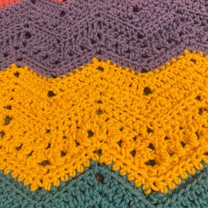 May include: A close-up of a handmade crochet blanket with a chevron pattern. The blanket features rows of coral, purple, yellow, and teal yarn. The crochet stitches create a textured surface with small openings throughout the design.