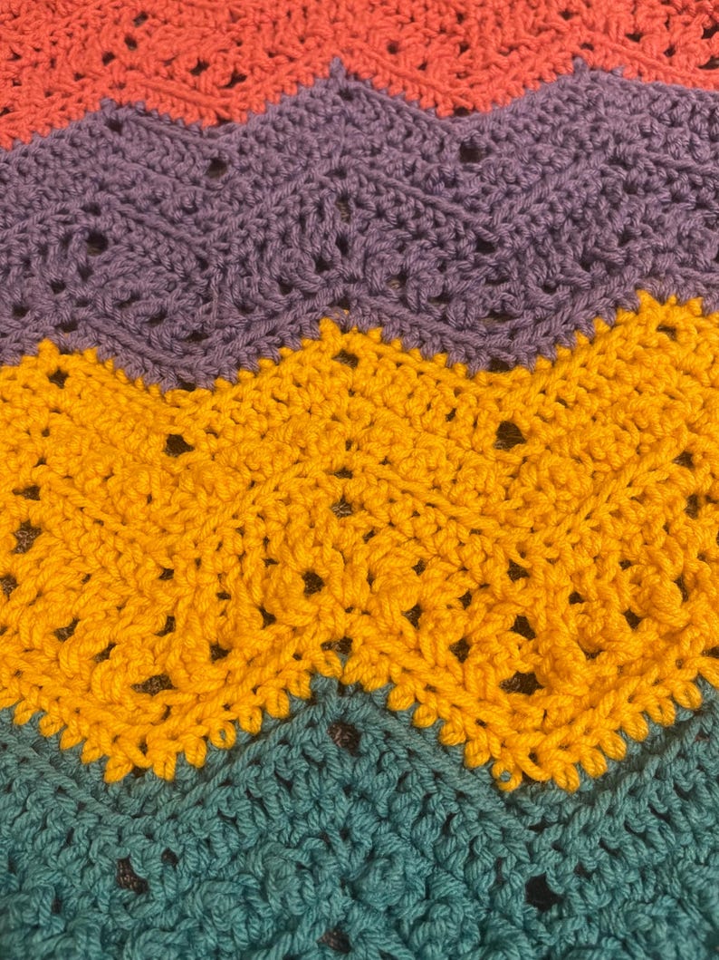May include: A close-up of a handmade crochet blanket with a chevron pattern. The blanket features rows of coral, purple, yellow, and teal yarn. The crochet stitches create a textured surface with small openings throughout the design.