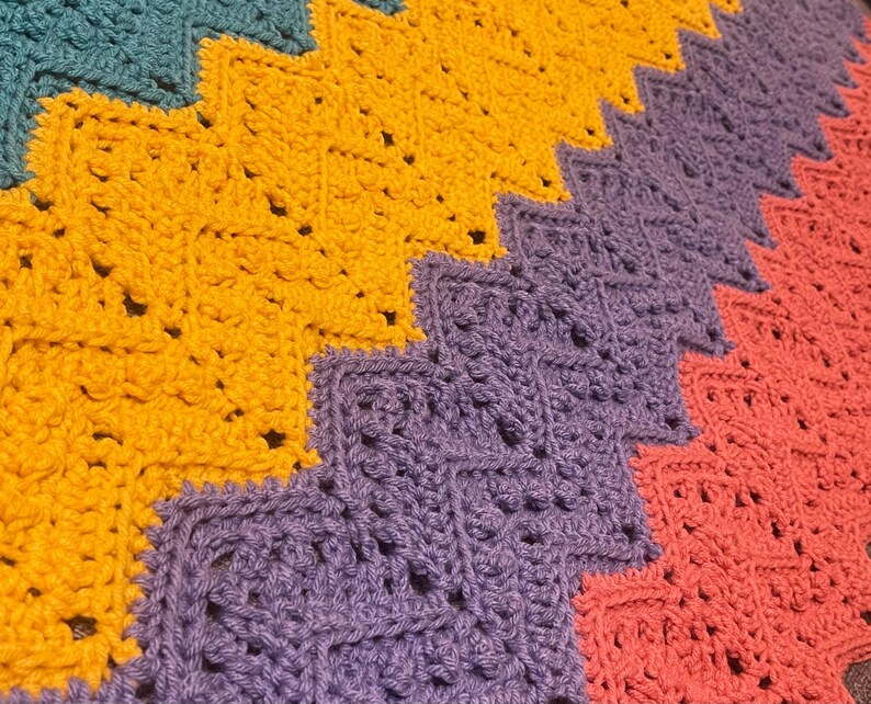 May include: Close-up of a handmade crochet blanket with a zig-zag pattern. The blanket features sections of teal, gold, purple, and coral. The stitches are visible, creating a textured surface. This cozy blanket is perfect for home decor.